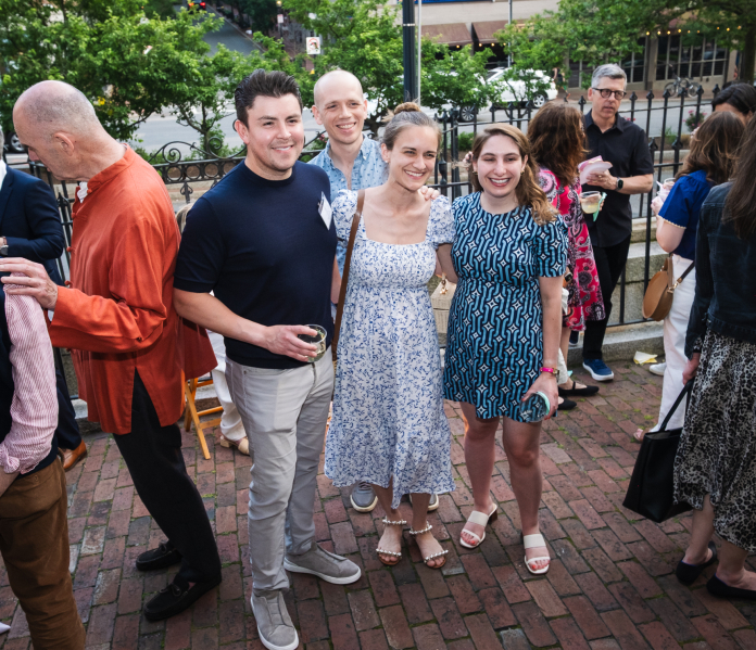 Four individuals face the camera, surrounded by many others who are not the focus of this image. They are standing on a red brick terrace with a black cast iron fence and green trees in the background.